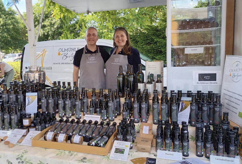 Zwei Personen stehen hinter einem Marktstand, an dem auf Handwerkermärkten und Bauernmärkten verschiedene Ölflaschen mit der Aufschrift "Ölmühle Bröker" und Flaschen mit verschiedenen Ölsorten des Spätsommer Herbst 2022 angeboten werden.