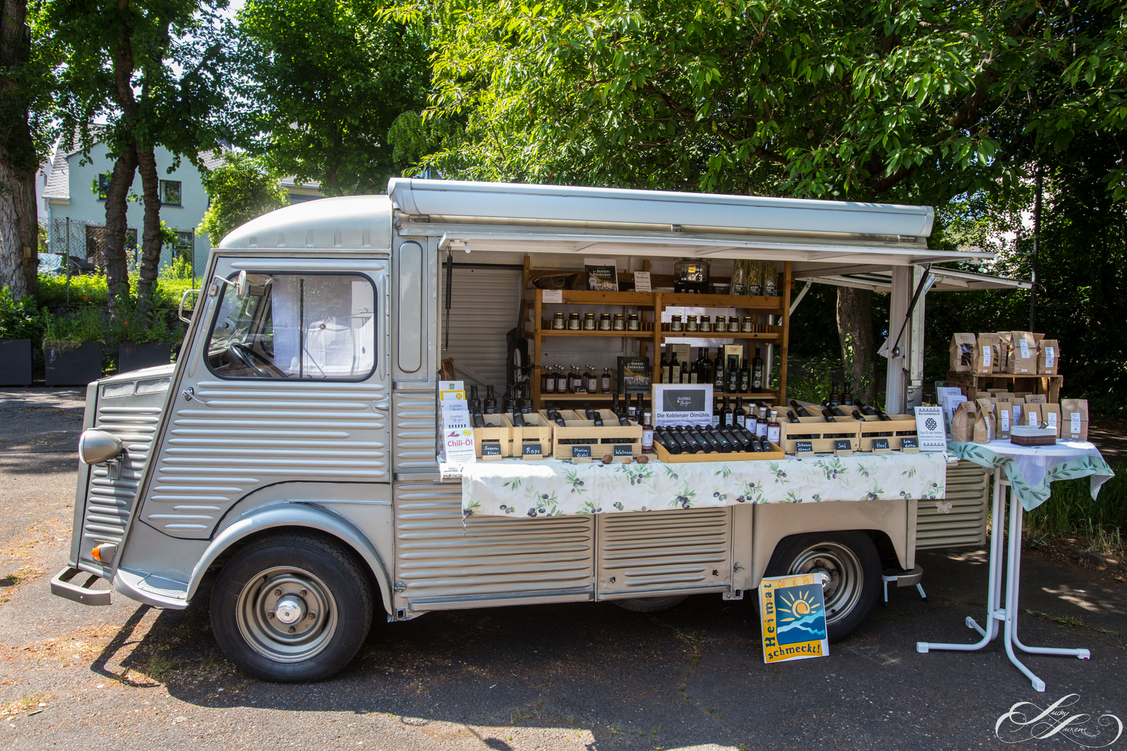 Ein silberner Foodtruck auf dem Wochenmarkt Ehrenbreitstein stellt verschiedene Flaschen und Gläser auf Regalen und einem Tisch im Freien aus, umgeben von Bäumen.