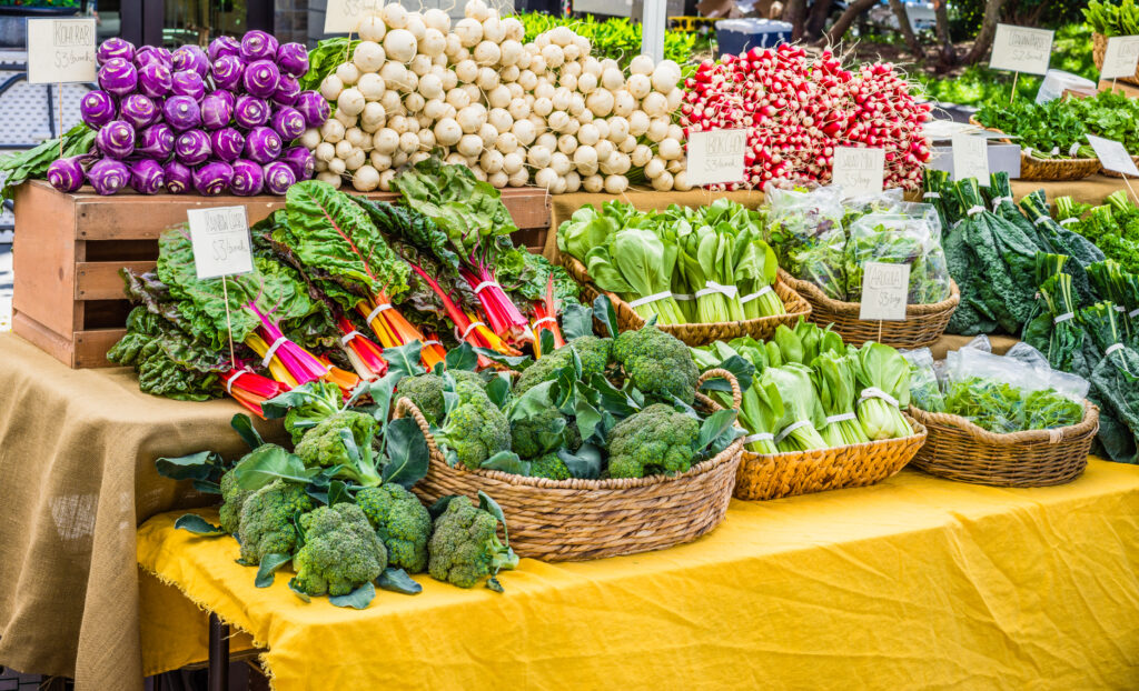 Auf dem Wochenmarkt Güls werden an einem Bauernmarktstand verschiedene frische Gemüsesorten wie Brokkoli, Mangold, Rüben, Radieschen, Grünkohl und Tütengemüse auf einer gelben Tischdecke angeboten.