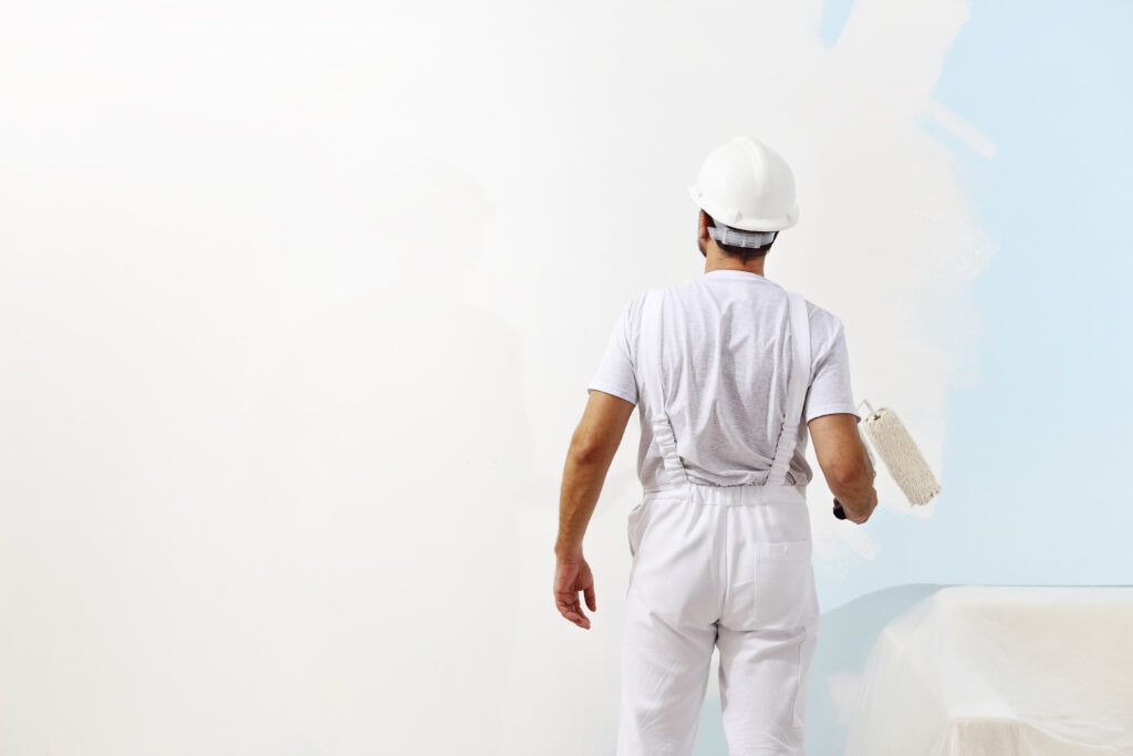 Eine Person, die einen weißen Overall und einen Schutzhelm trägt, streicht mit einer Walze eine Wand weiß, während sie von der Kamera wegschaut.